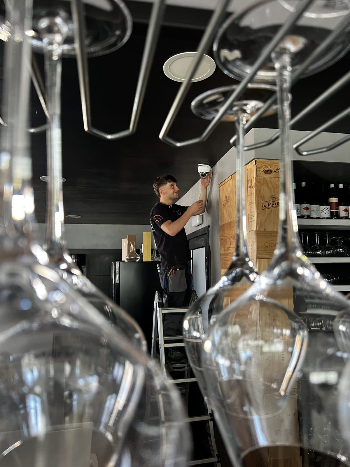 A Buzz Alarmas engineer fitting a camera inside a restaurant, seen through hanging wine glasses.